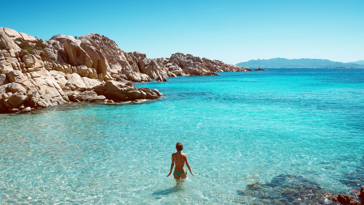 En kvinna i bikini vadar ut i klart turkost vatten vid en stenig strand.