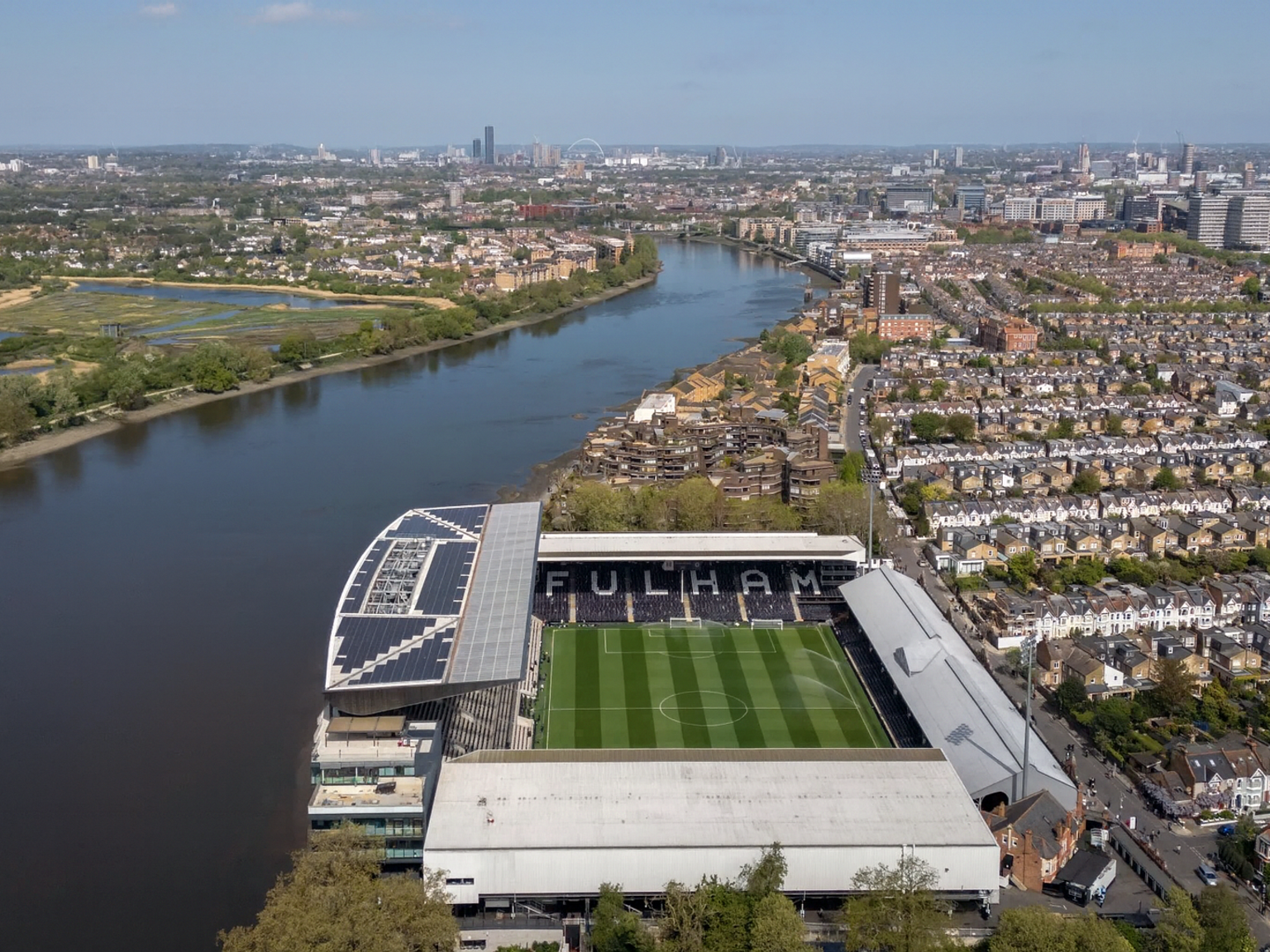 Luchtfoto van het stadion van Fulham Football Club aan de Theems, omringd door gebouwen en een verre stadsskyline.