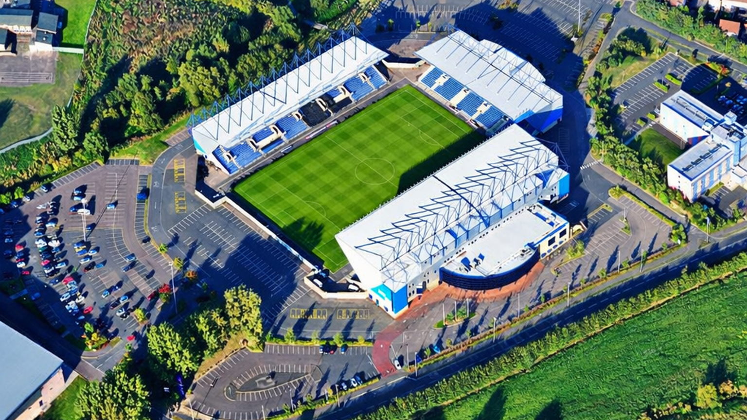 Luftaufnahme eines Fußballstadions mit einem grünen Spielfeld, umgeben von Parkplätzen, Bäumen und Wohngebieten.