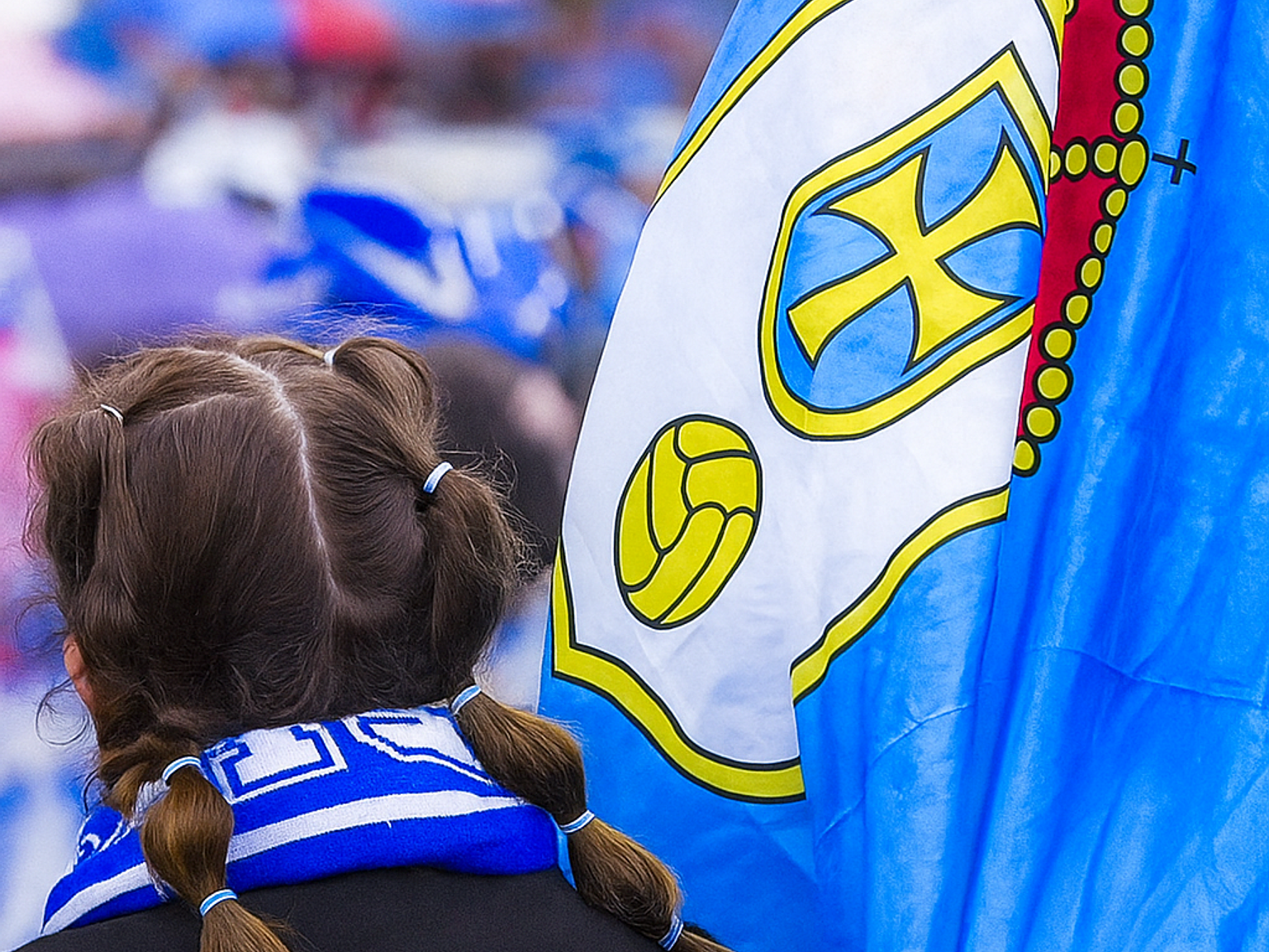Rückansicht einer Person mit Zöpfen und einem Oviedo-Schal neben einer blau-weißen Flagge mit einem Wappen.