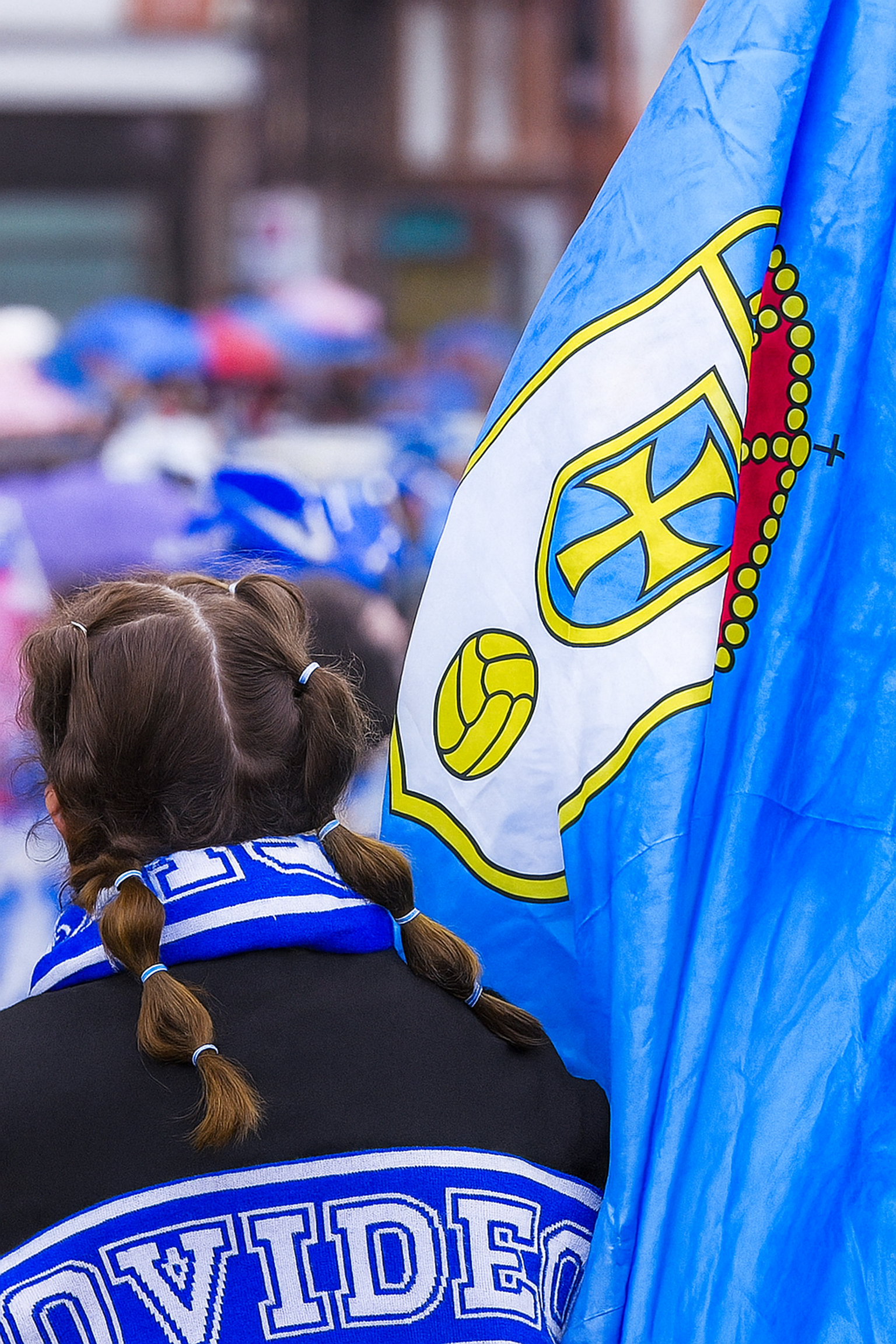 Rückansicht einer Person mit Zöpfen und einem Oviedo-Schal neben einer blau-weißen Flagge mit einem Wappen.