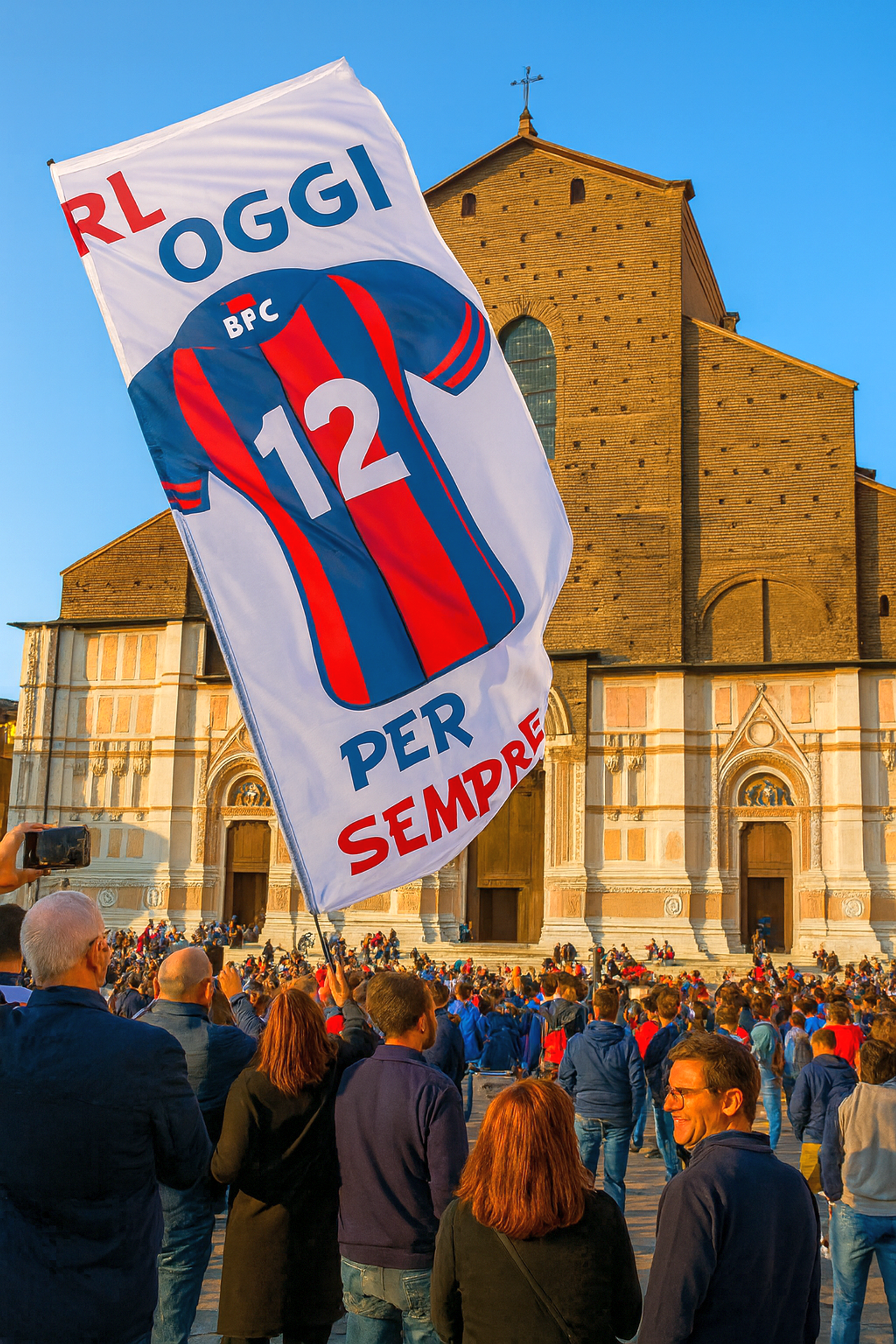 Un grand drapeau avec un maillot de football rayé rouge et bleu (numéro 12) et le texte italien "OGGI PER SEMPRE" flotte au-dessus d'une foule devant une cathédrale en brique.