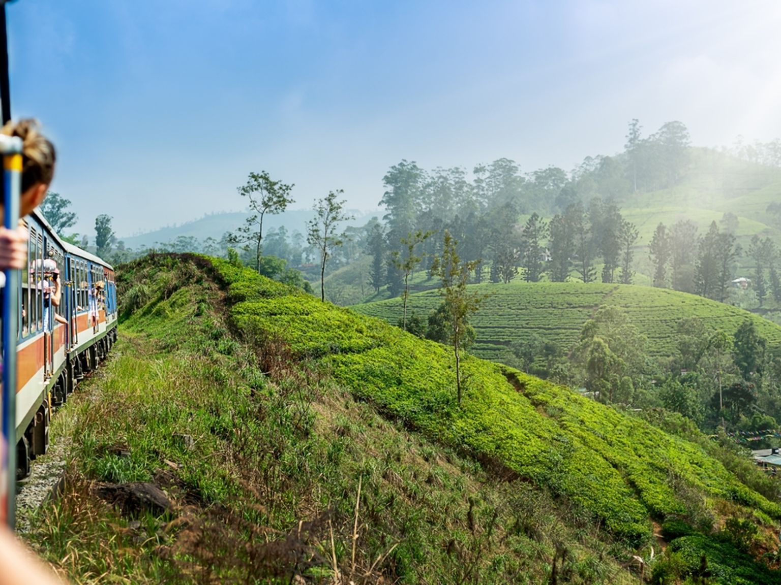 Passagerare tittar ut från ett tåg som slingrar sig genom frodiga gröna teplantager på böljande kullar. Sri Lanka