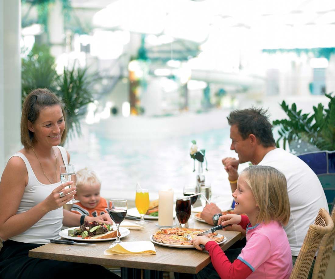En leende familj på fyra äter middag vid ett restaurangbord med en simbassäng synlig i bakgrunden.