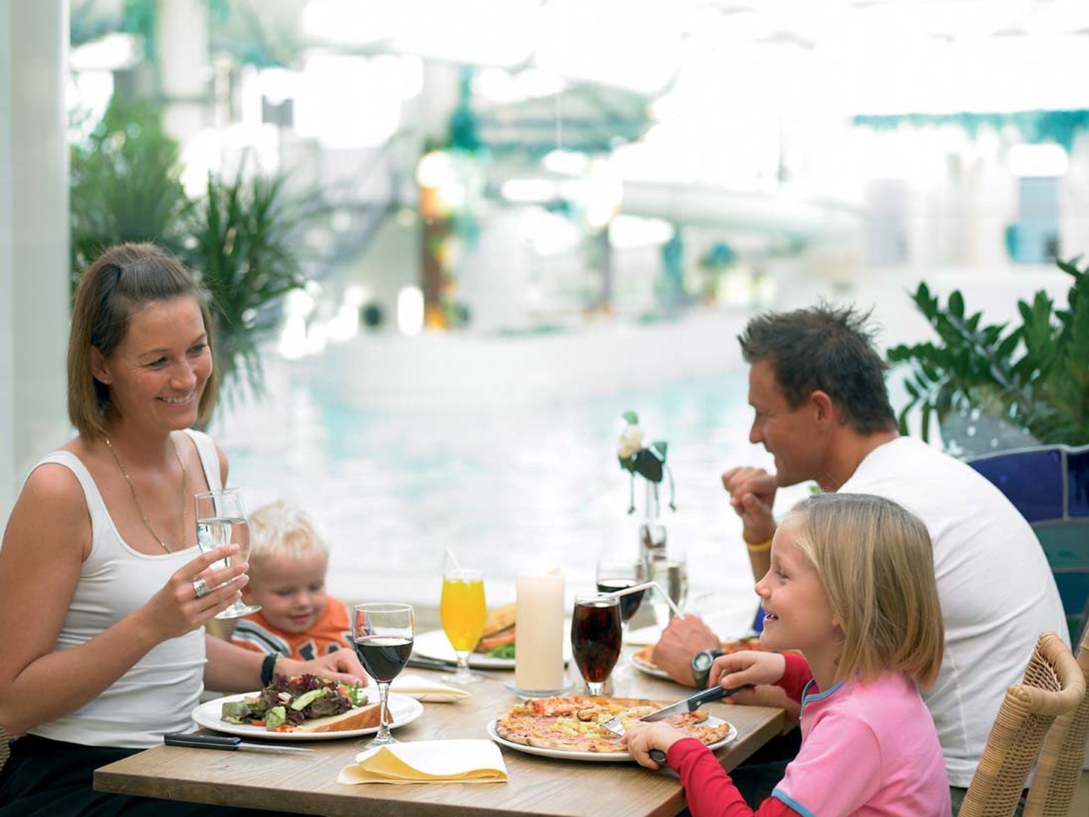 En leende familj på fyra äter middag vid ett restaurangbord med en simbassäng synlig i bakgrunden.