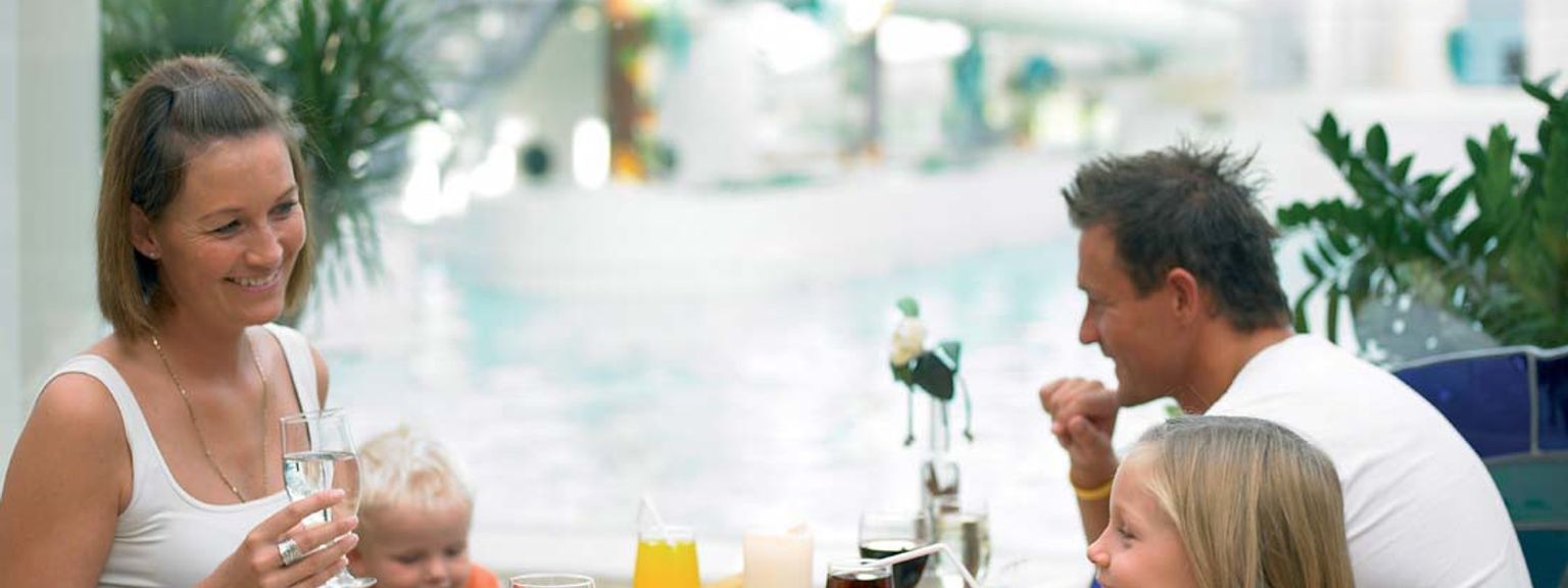 En leende familj på fyra äter middag vid ett restaurangbord med en simbassäng synlig i bakgrunden.