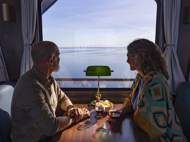 En man och en kvinna i en tåg- eller båthytt som njuter av vin och snacks medan de tittar ut på en havsbaserad vindkraftpark.