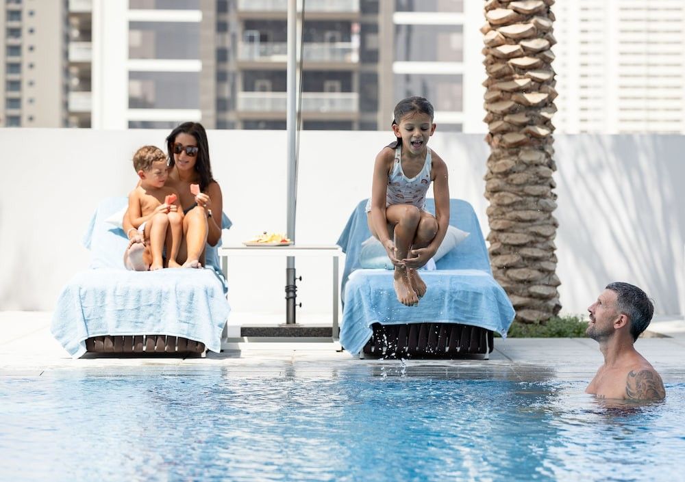 En familie ved et basseng. En kvinne og en gutt spiser ispinner på en solseng mens en jente hopper i vannet, mens en mann ser på fra bassenget.