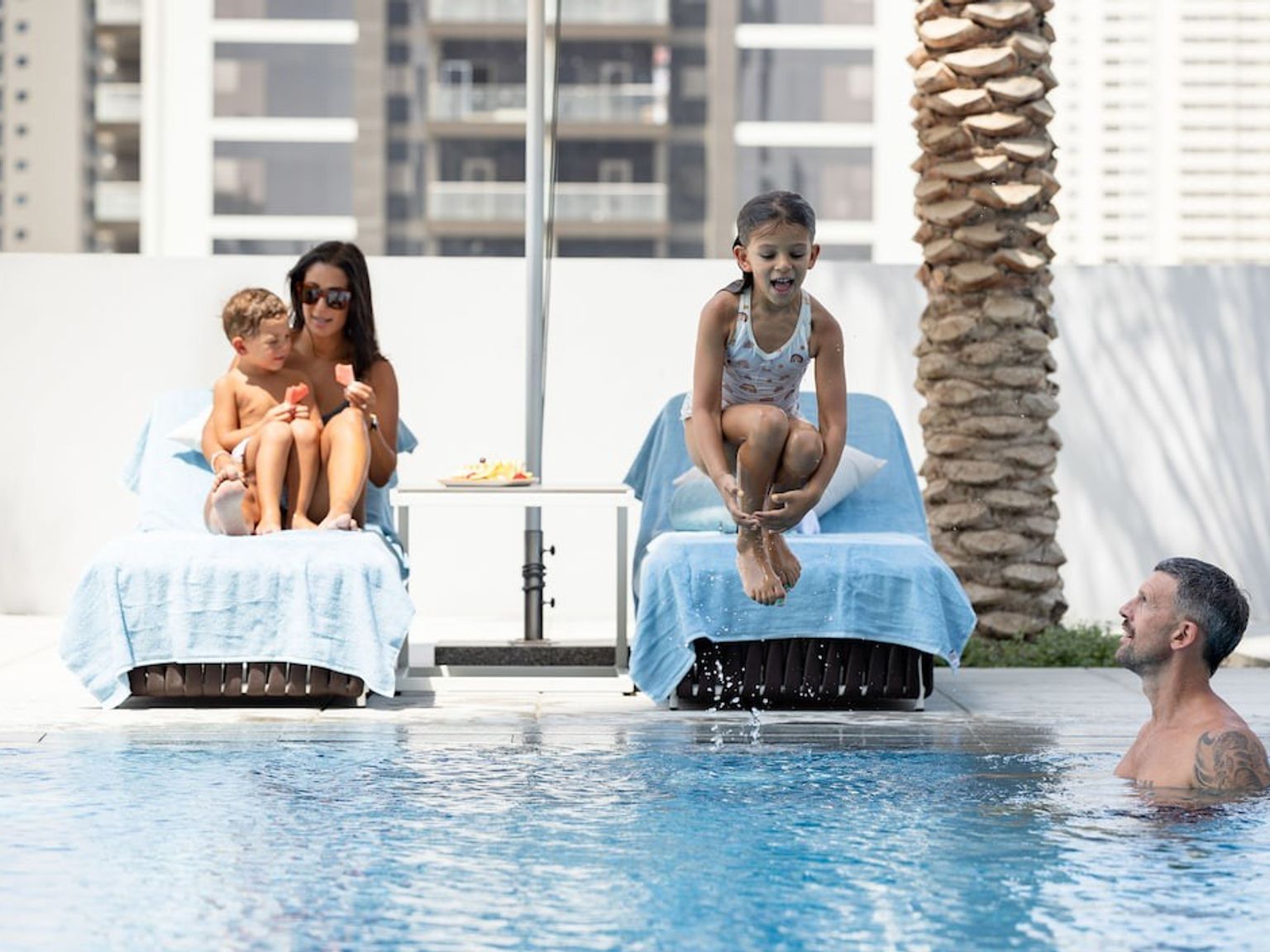 En familie ved et basseng. En kvinne og en gutt spiser ispinner på en solseng mens en jente hopper i vannet, mens en mann ser på fra bassenget.