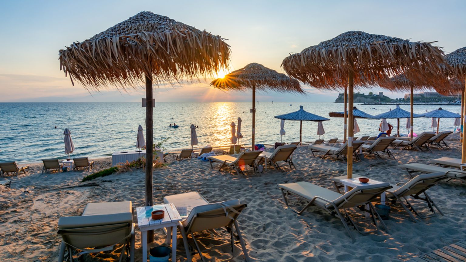 En sandstrand ved solnedgang med stråparasoller og liggestole med udsigt over havet.