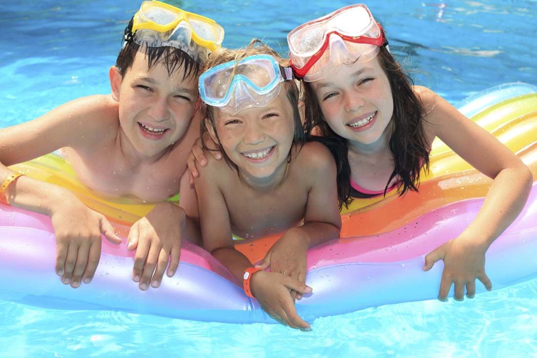 Tre smilende barn med svømmebriller på hodet flyter på en fargerik flåte i et basseng.