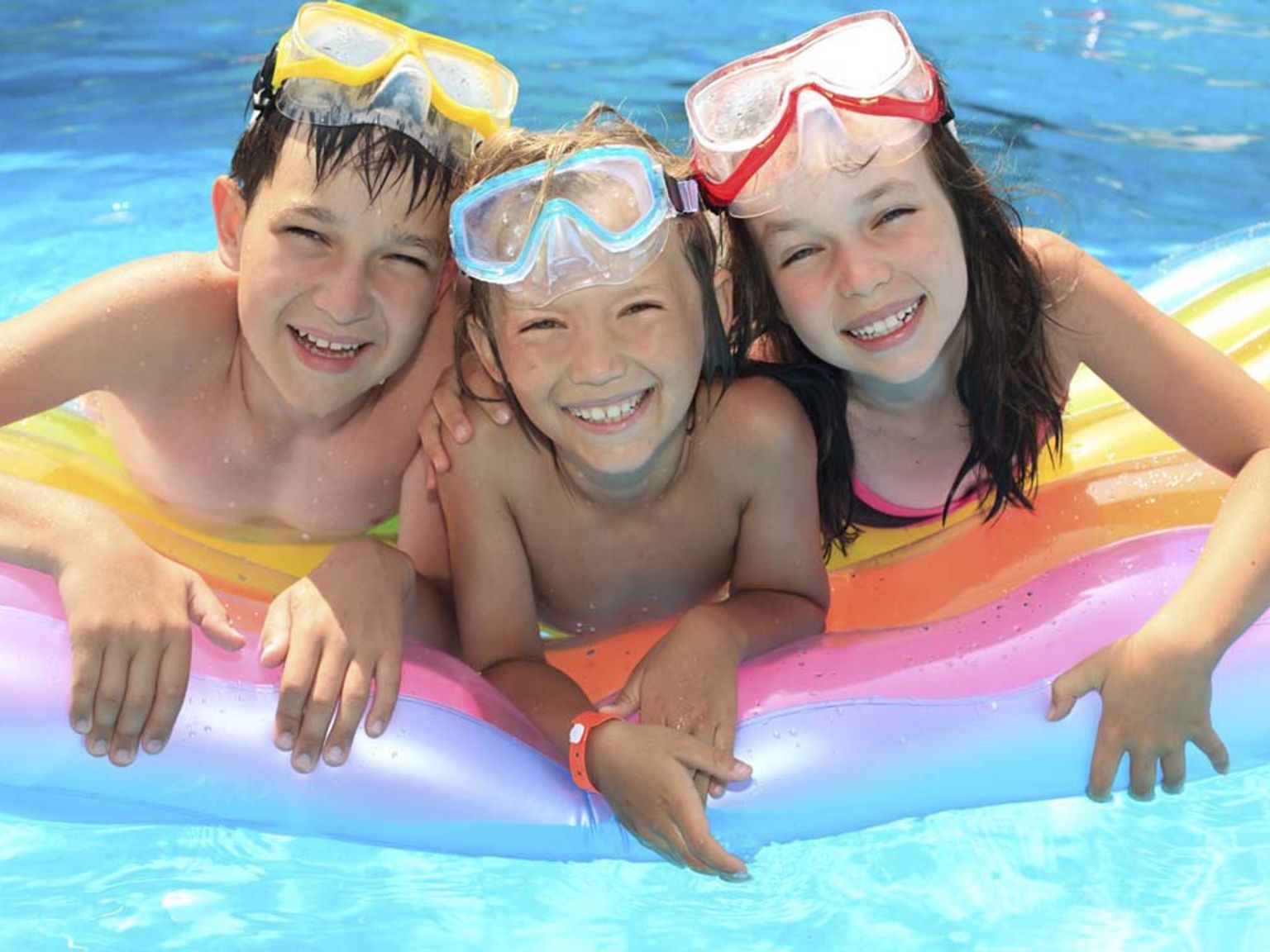 Tre smilende barn med svømmebriller på hodet flyter på en fargerik flåte i et basseng.