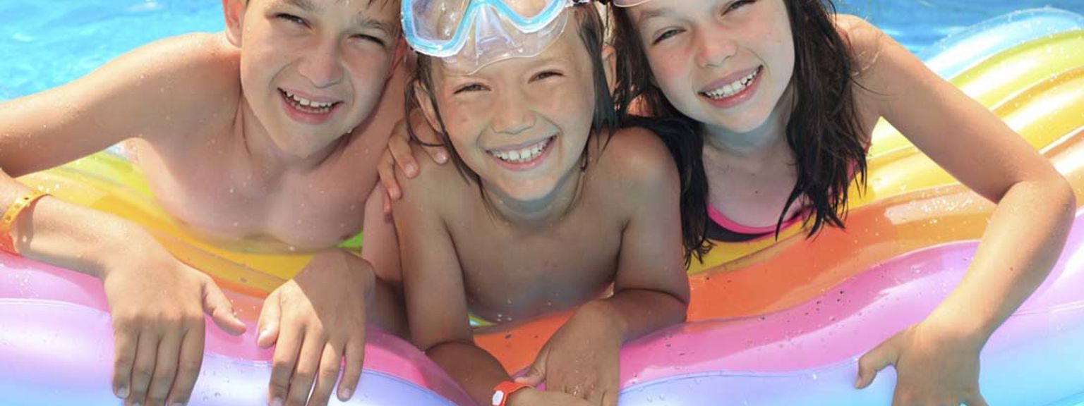 Tre smilende barn med svømmebriller på hodet flyter på en fargerik flåte i et basseng.
