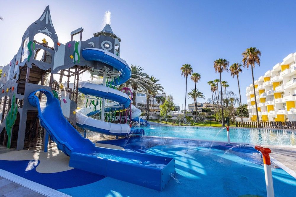 Slot-tema vandland med rutsjebaner og plaskebassin ved siden af en stor pool, palmer og feriested bygninger.