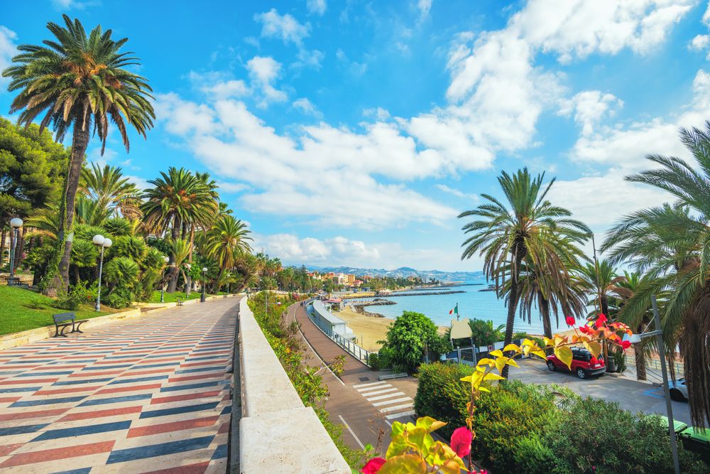En mønstret promenade med palmer langs siderne har udsigt over en sandstrand og et blåt hav under en klar himmel. San Remo
