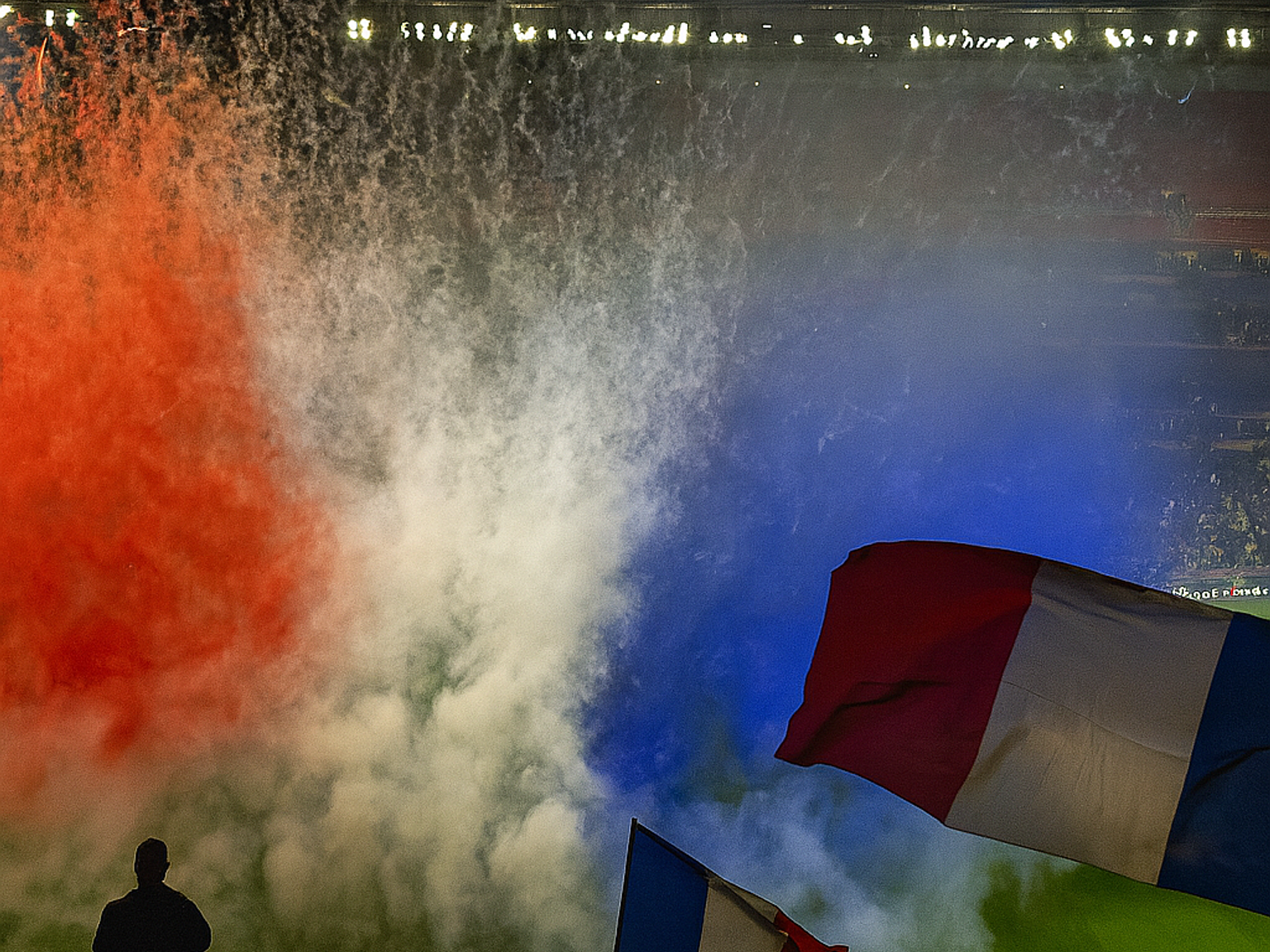 Roter, weißer und blauer Rauch füllt ein Stadion, während eine silhouettierte Menge französische Flaggen unter einem UEFA-Banner schwenkt.