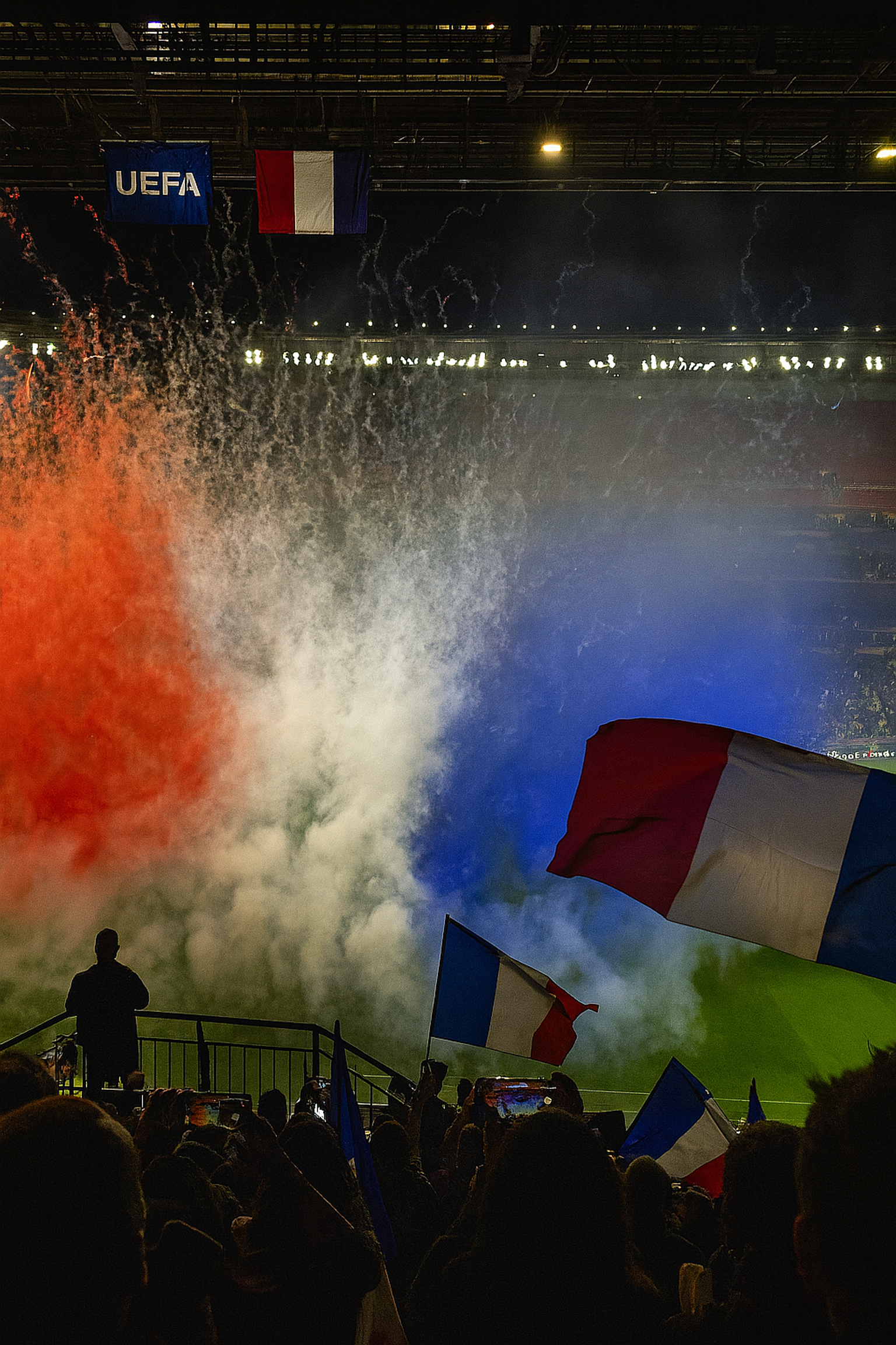 Roter, weißer und blauer Rauch füllt ein Stadion, während eine silhouettierte Menge französische Flaggen unter einem UEFA-Banner schwenkt.