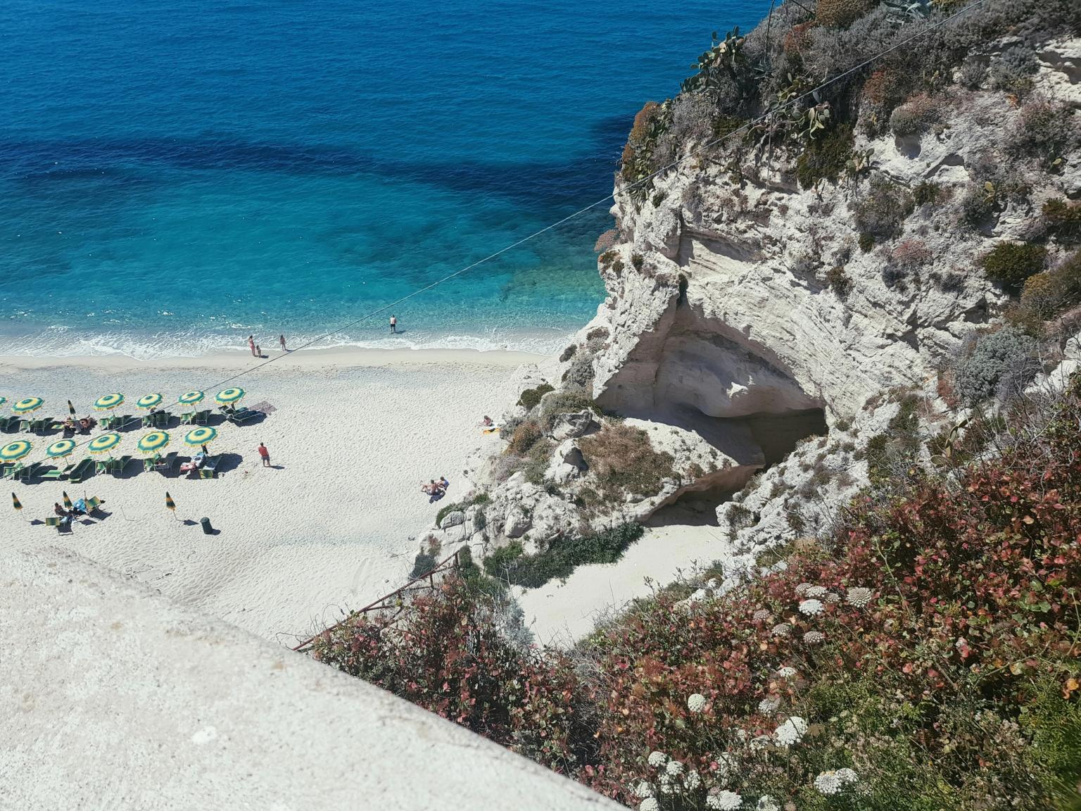 Vista dall’alto di una spiaggia di sabbia bianca con ombrelloni verdi, mare turchese e una scogliera rocciosa con una grotta. Tropea, Calabria