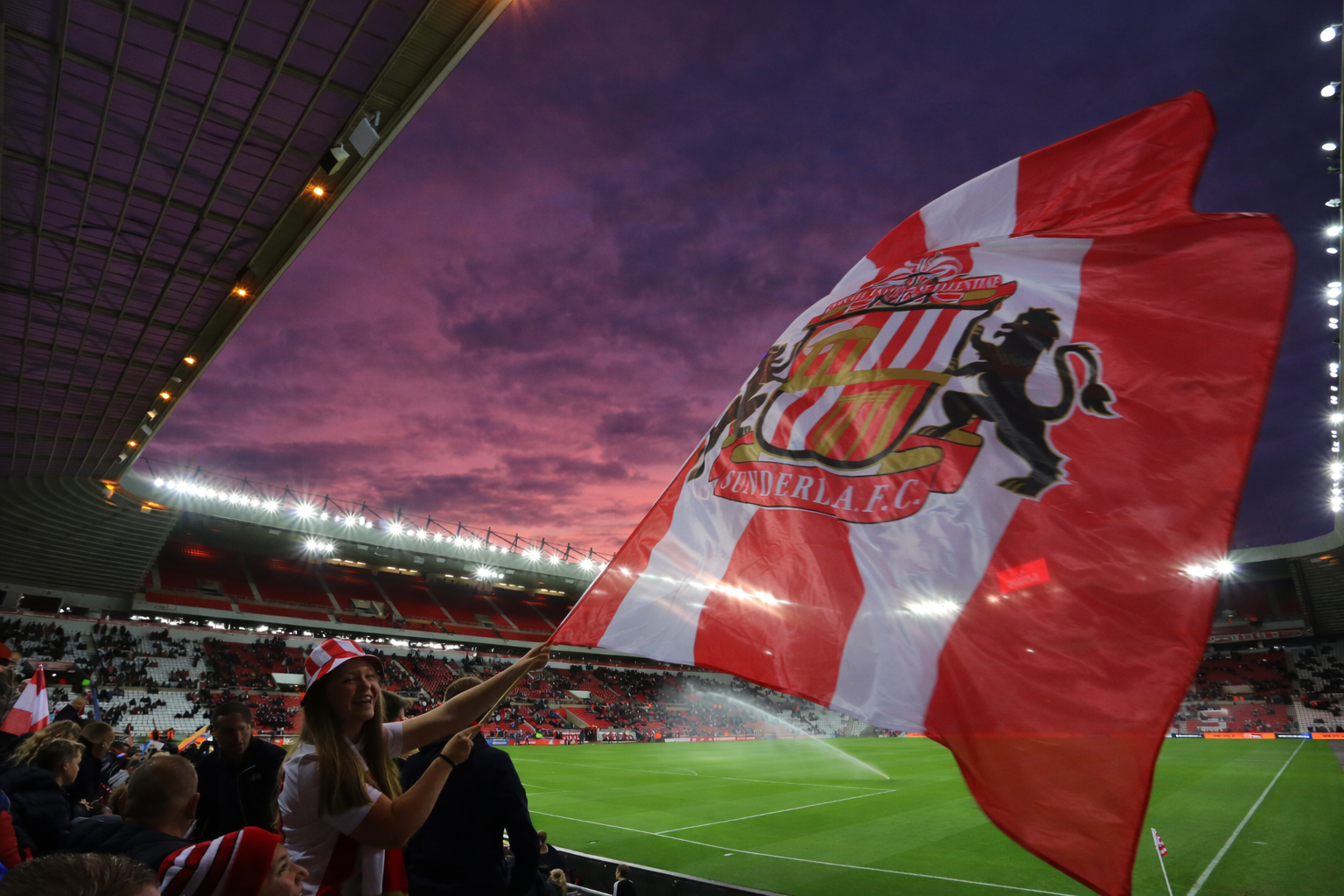 A fan waves a red and white Sunderland F.C. flag against a dramatic purple sunset over a football stadium.