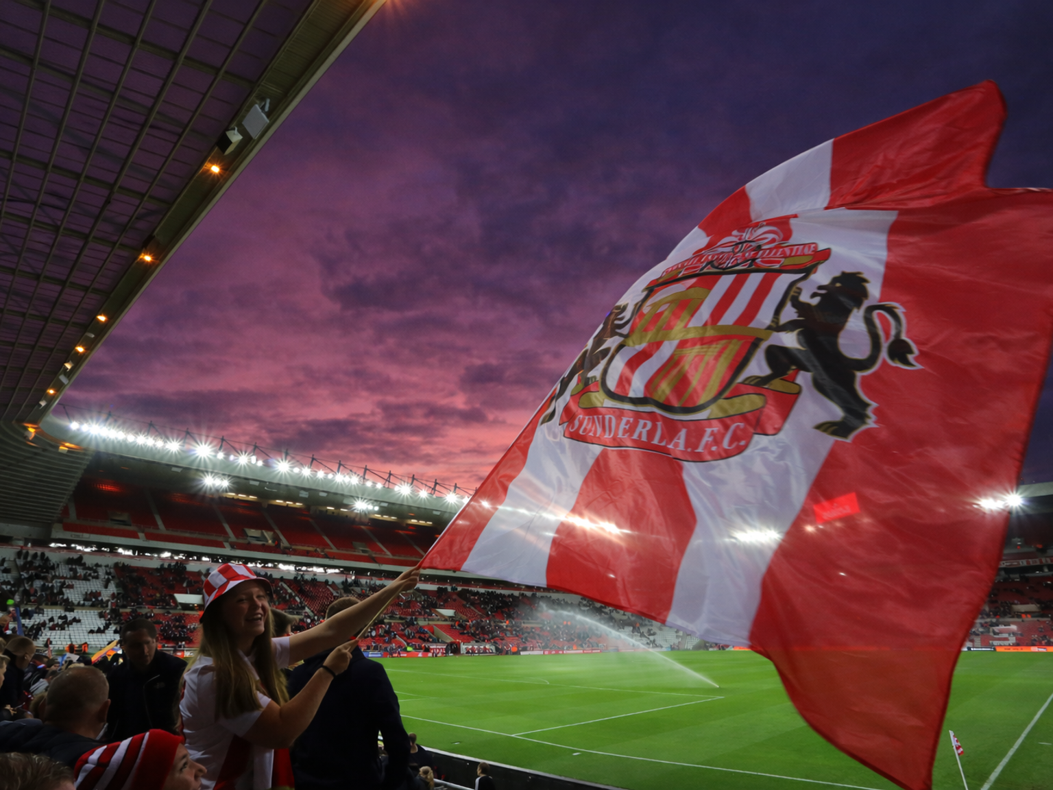 A fan waves a red and white Sunderland F.C. flag against a dramatic purple sunset over a football stadium.