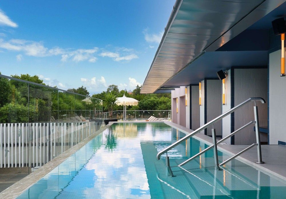 Modern utomhus-infinitypool som speglar den blå himlen, flankerad av en samtida byggnad och gröna träd.