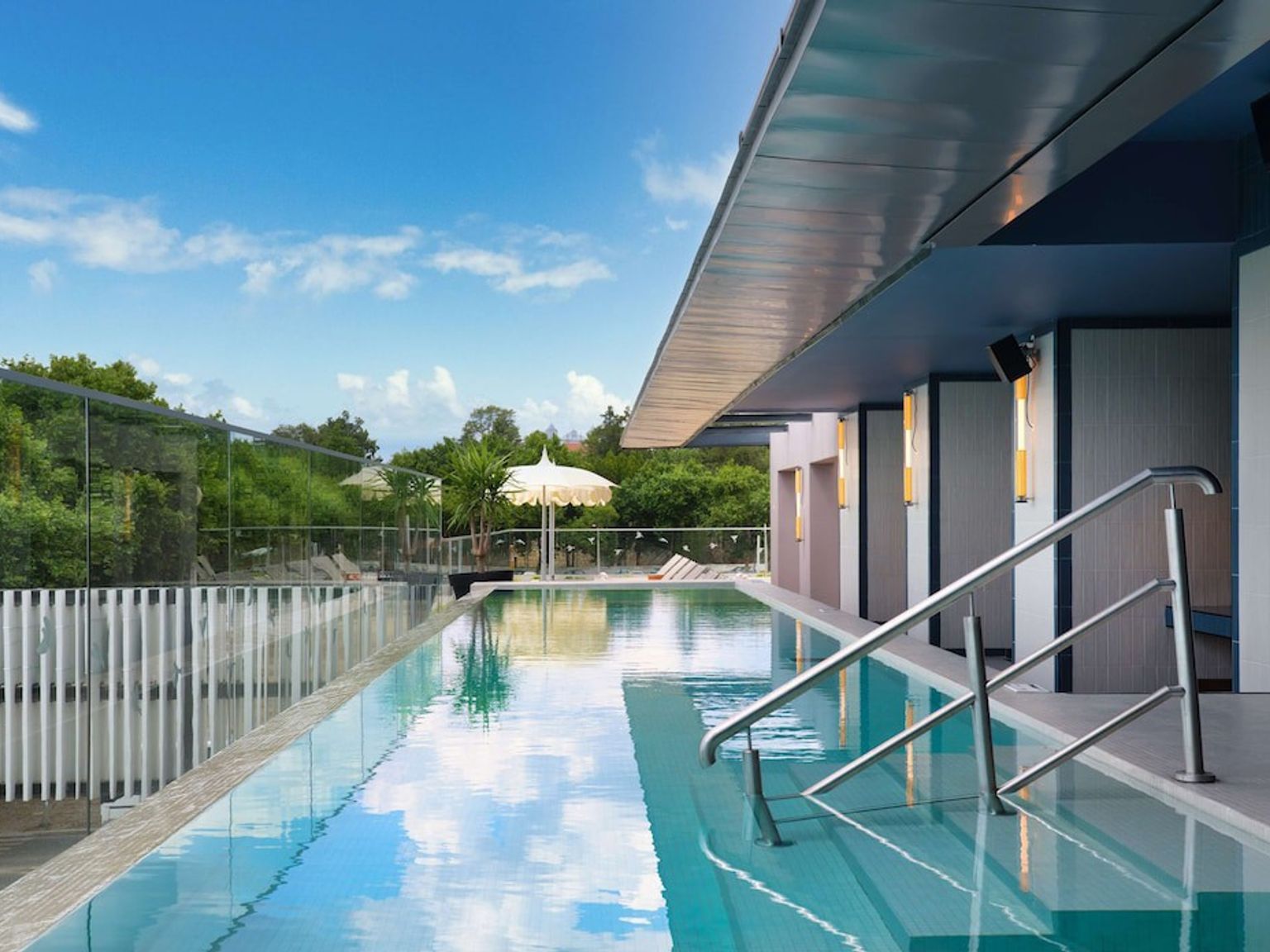 Modern utomhus-infinitypool som speglar den blå himlen, flankerad av en samtida byggnad och gröna träd.