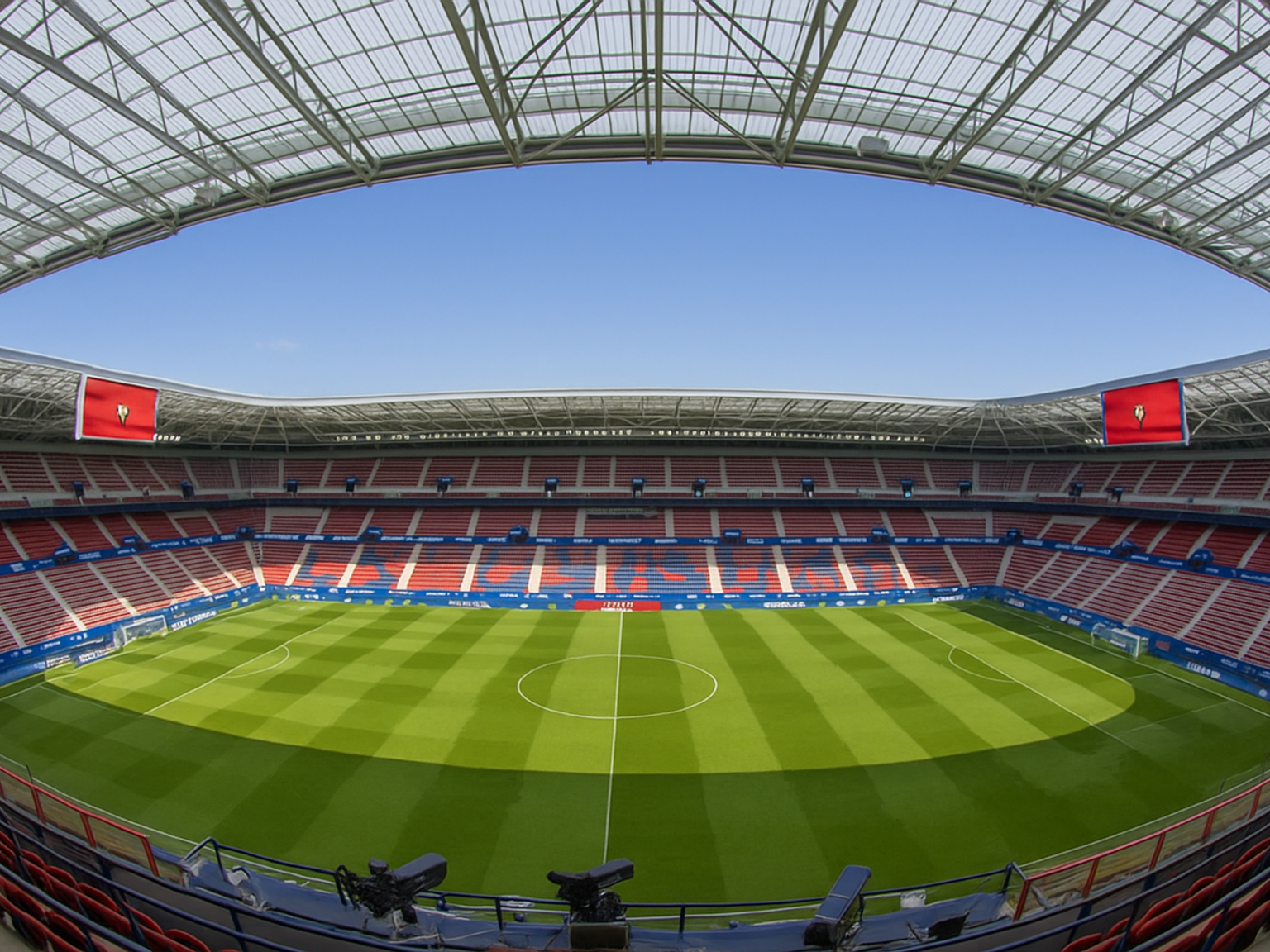 Leeg modern voetbalstadion met een groen veld, rode en blauwe stoelen en een doorschijnend dak onder een blauwe lucht.