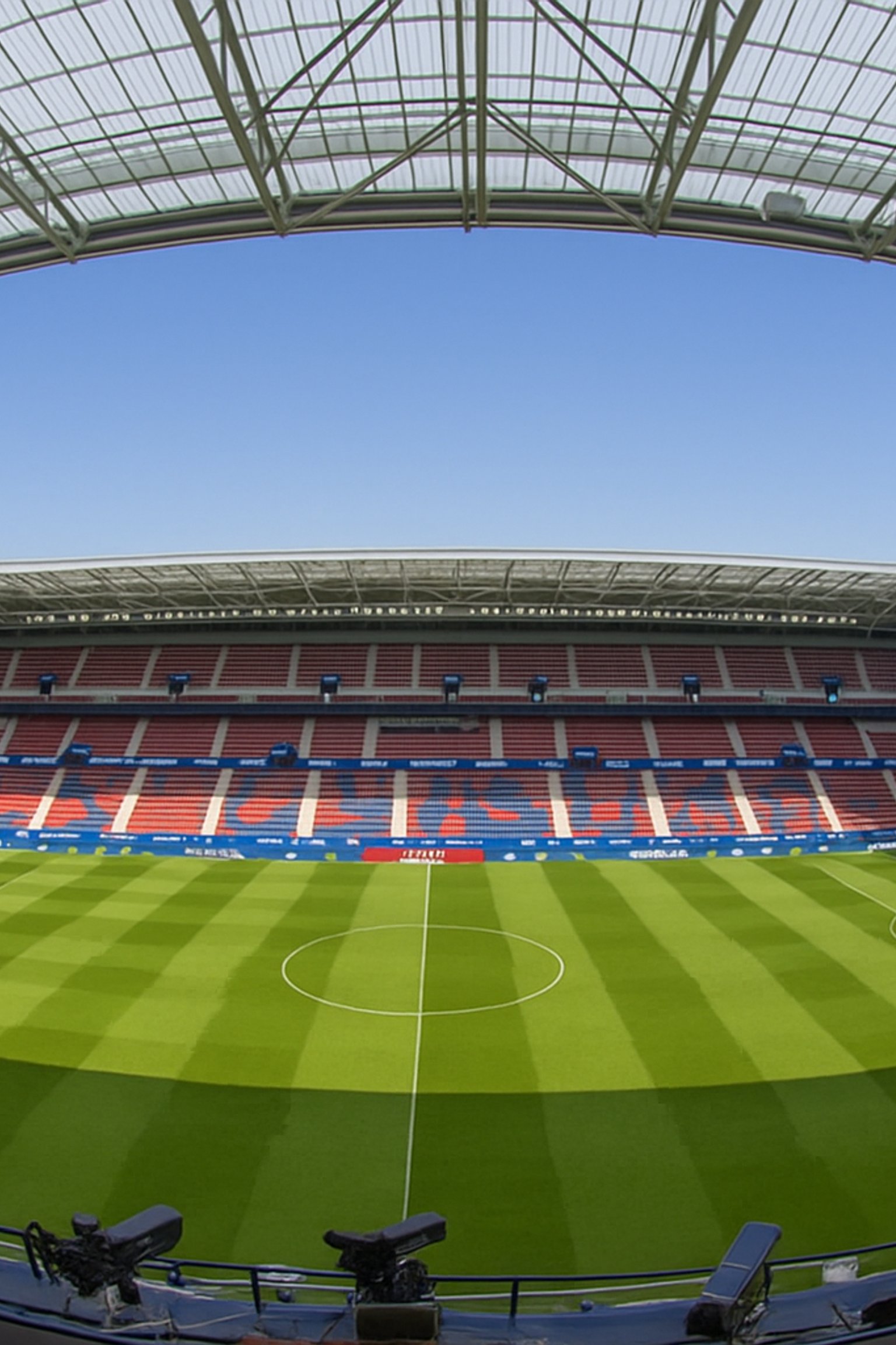 Leeg modern voetbalstadion met een groen veld, rode en blauwe stoelen en een doorschijnend dak onder een blauwe lucht.