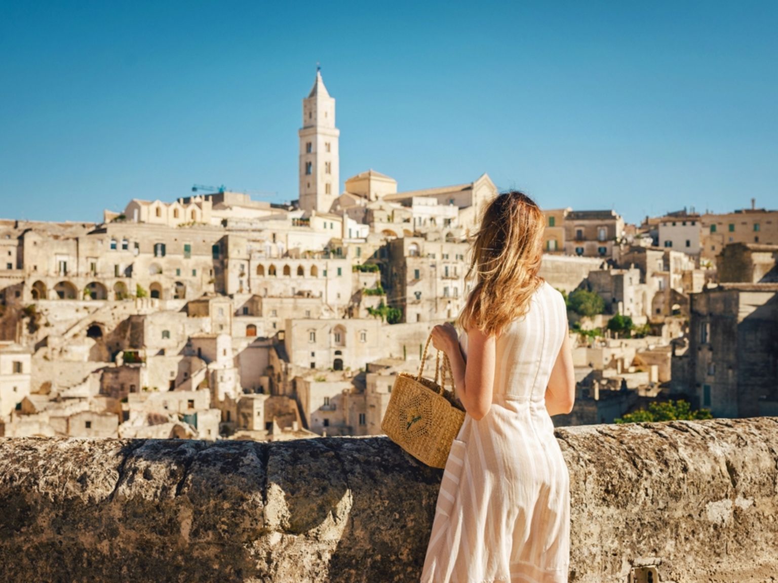 Kvinne ser ut over en gammel by med steinbygninger og et høyt klokketårn under en klar blå himmel. Matera, Italia
