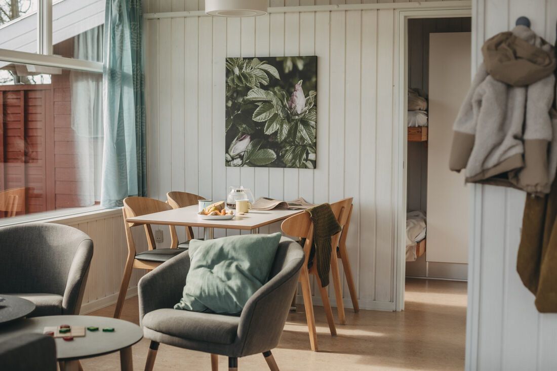 Ett ljust, mysigt vardagsrum och matplats med ett frukostbord, fåtöljer, ett stort fönster och en glimt in i ett sovrum med våningssäng.