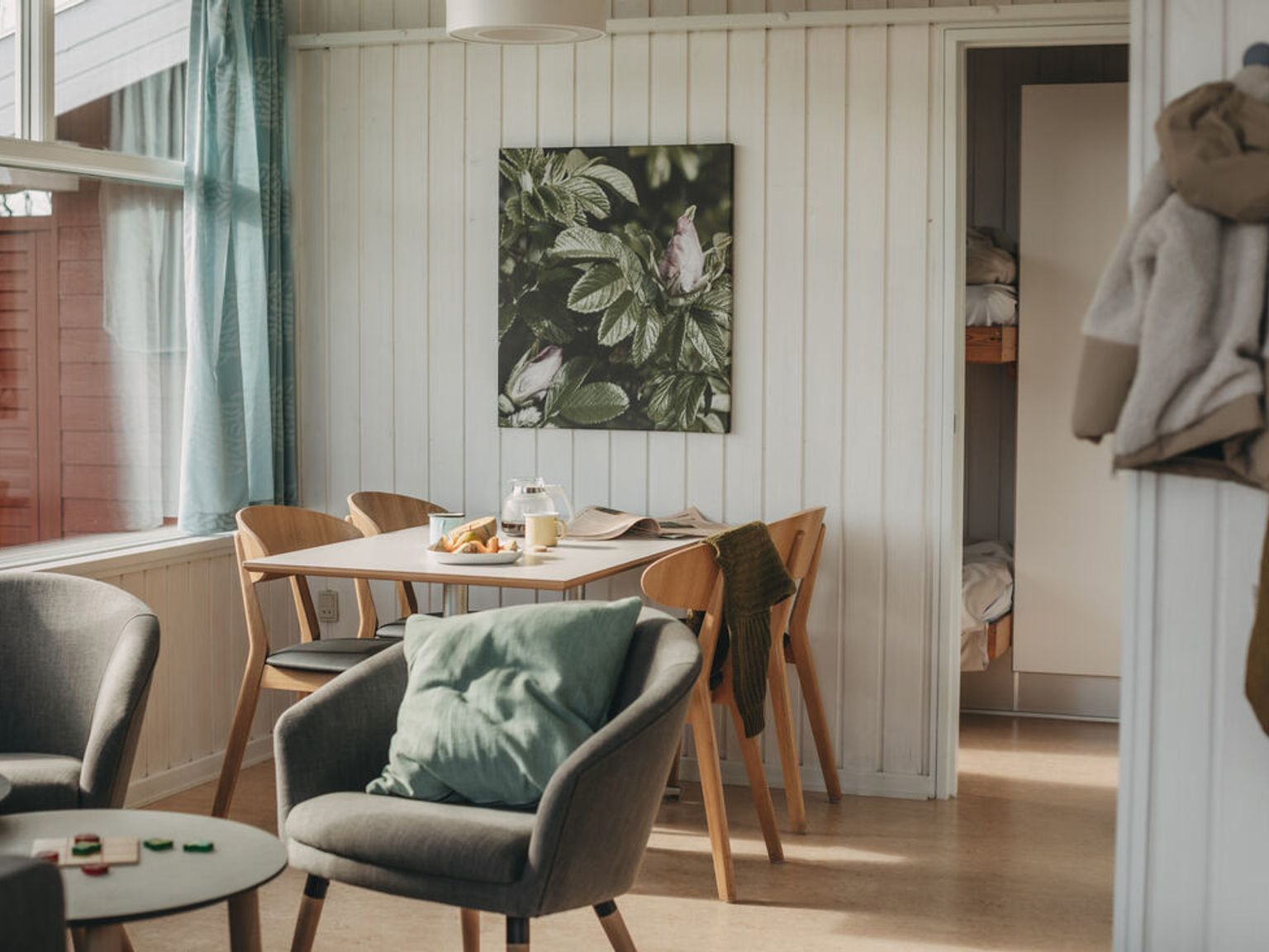 Et lyst, koselig stue- og spiseområde med et frokostbord, lenestoler, et stort vindu og et glimt inn i et køyeseng soverom.