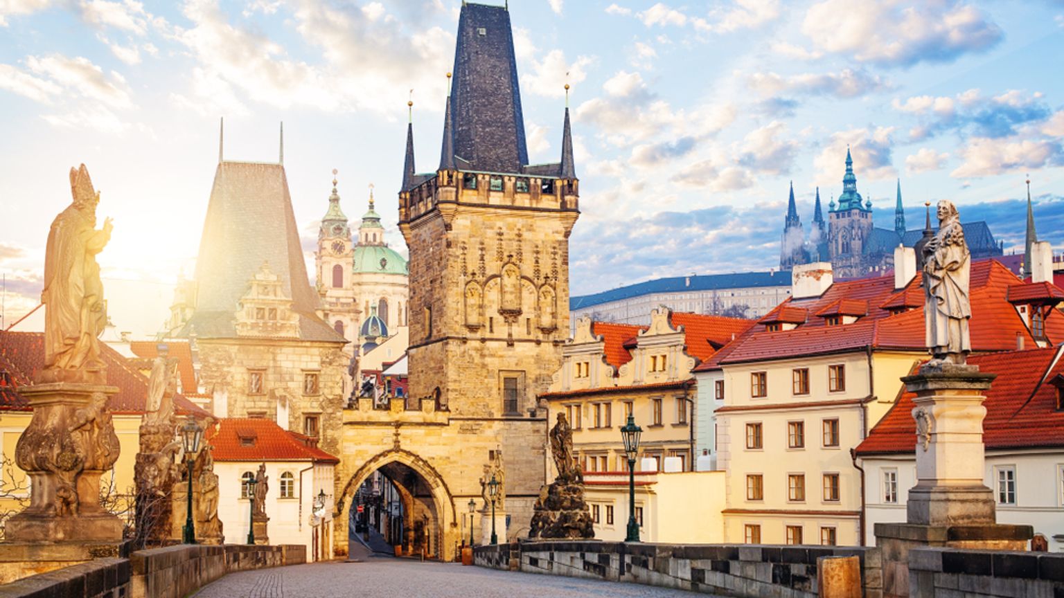Soluppgång på Karlsbron i Prag, med statyer, ett brotorn och ett avlägset slott.