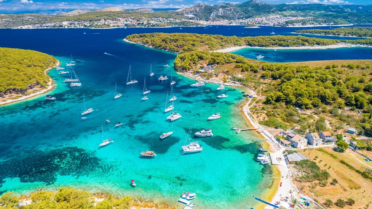 Luftfoto af en livlig turkis bugt fyldt med både, omgivet af frodige grønne øer og en fjern kystby. Hvar, Kroatien