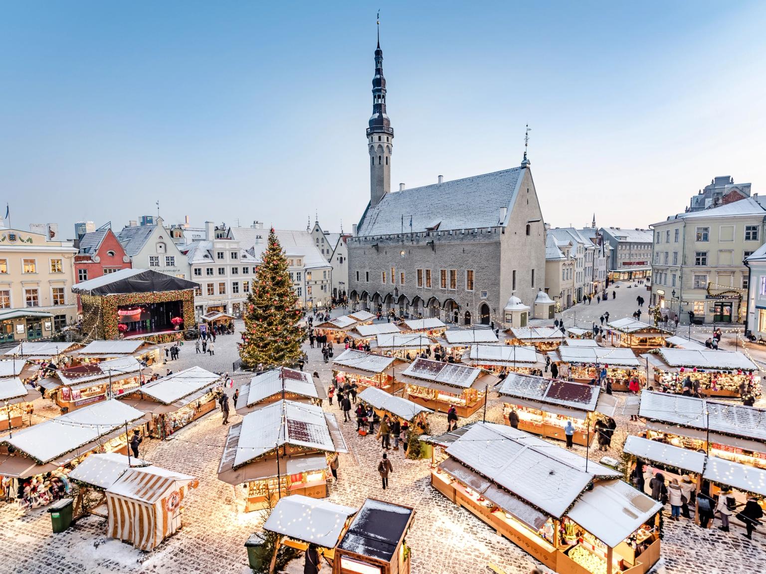 Et snødekt julemarked i Tallinn på et historisk torg med et stort dekorert tre og gamle bygninger.