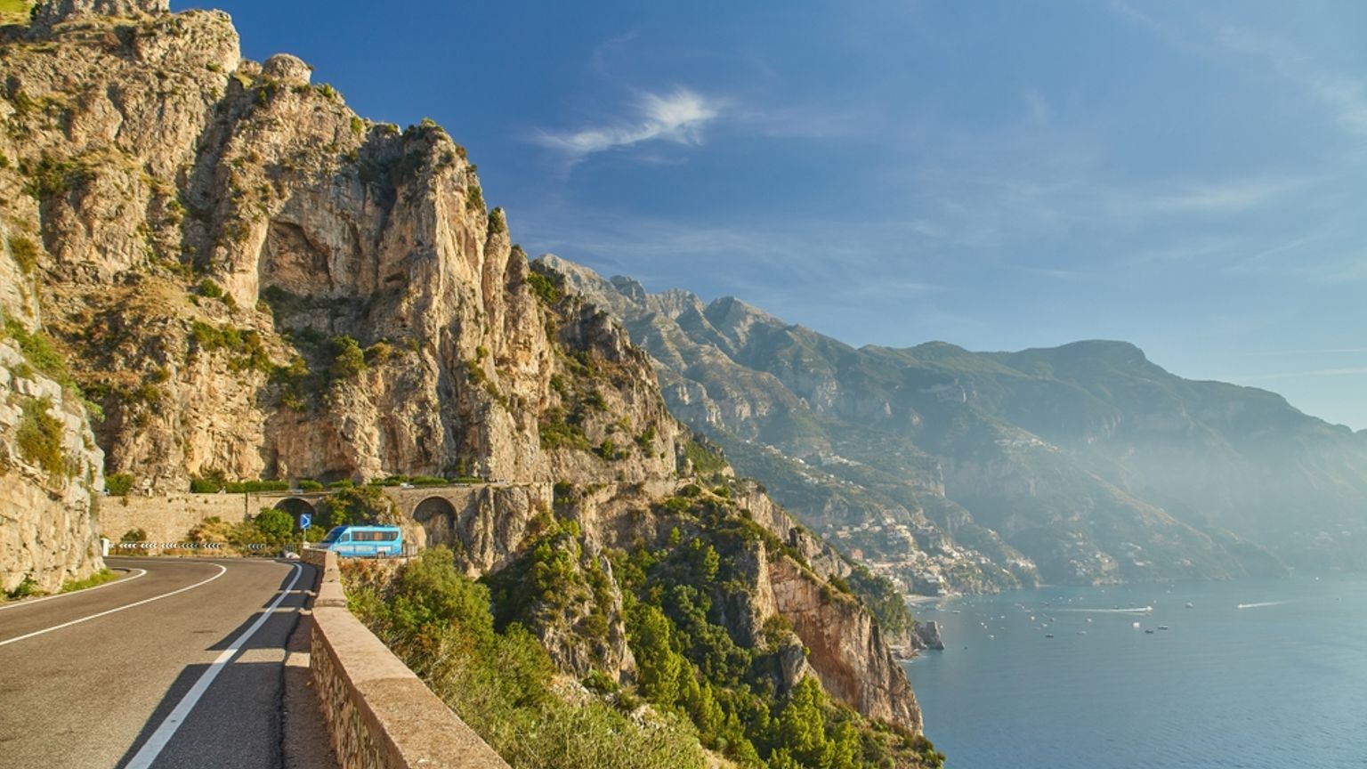 En slingrande kustväg med en blå buss som klamrar sig fast vid en brant, klippig klippsida med utsikt över det ljusblå havet.