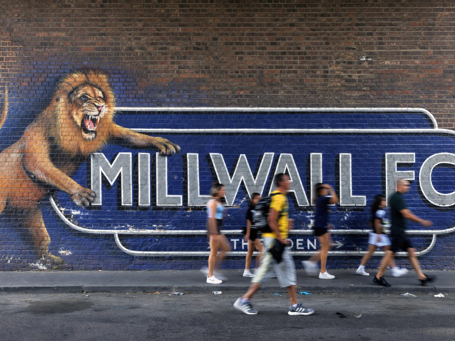 Mural de um leão rugindo e "MILLWALL FC" em uma parede de tijolos, com pessoas desfocadas passando.
