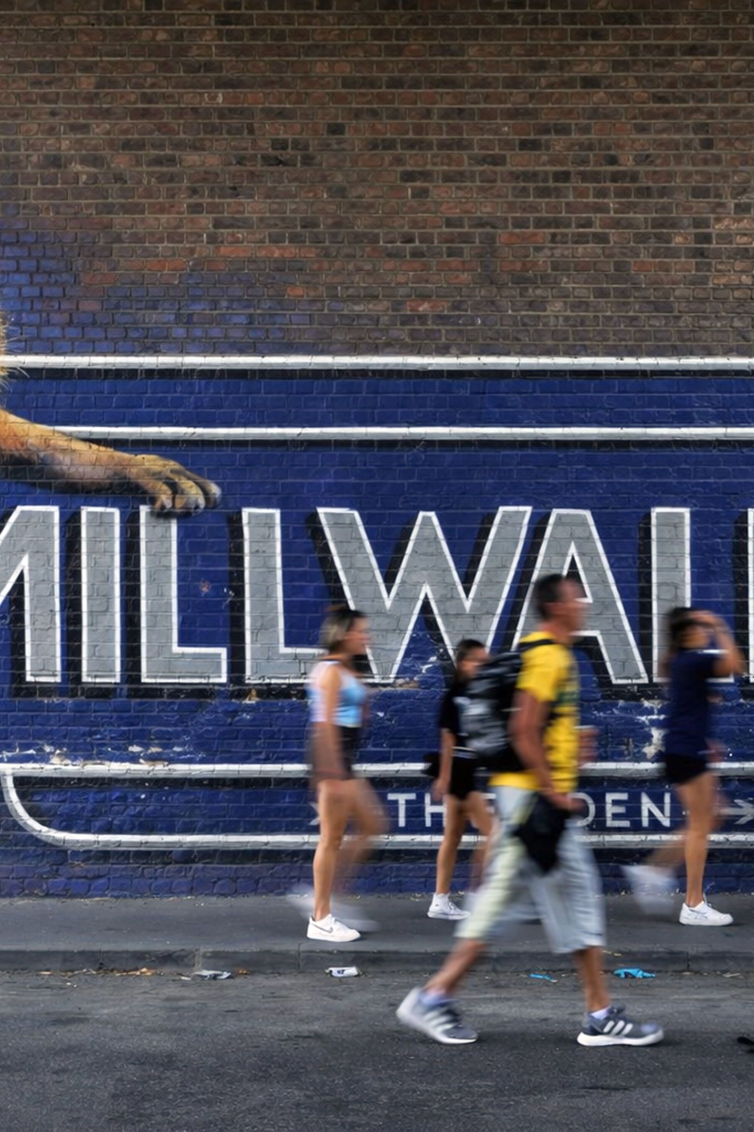 Mural de um leão rugindo e "MILLWALL FC" em uma parede de tijolos, com pessoas desfocadas passando.