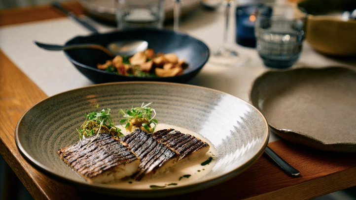 Tre grillede fiskefileter i en cremet sauce med grøn pynt på en mønstret tallerken ved et middagsbord.