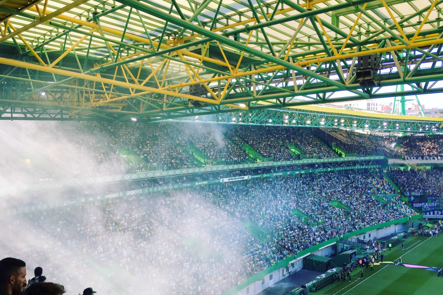 Packed stadium with a green and yellow roof, white smoke billowing over the stands filled with fans.