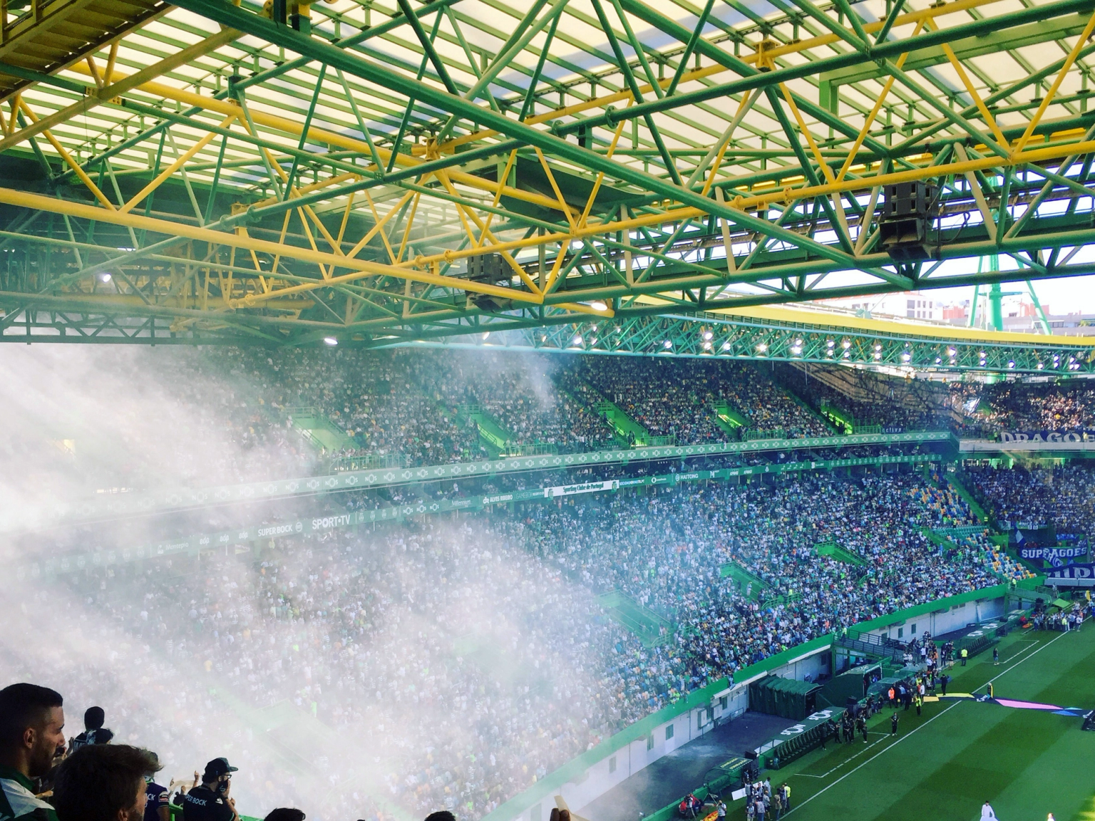 Packed stadium with a green and yellow roof, white smoke billowing over the stands filled with fans.