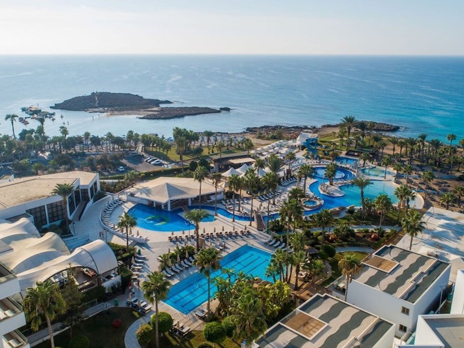 Luftfoto av et stort resortkompleks med flere blå bassenger, palmer og hvite bygninger langs en steinete kyst ved siden av et klart blått hav.