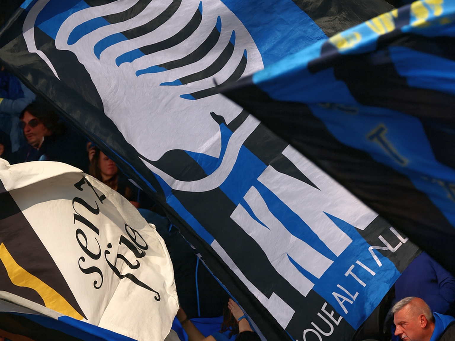 Jalkapallofanit stadionilla heiluttavat suuria mustia, sinisiä ja valkoisia lippuja, joista yhdessä on näkyvästi tyylitelty vaakuna ja sana "ATALANTA".