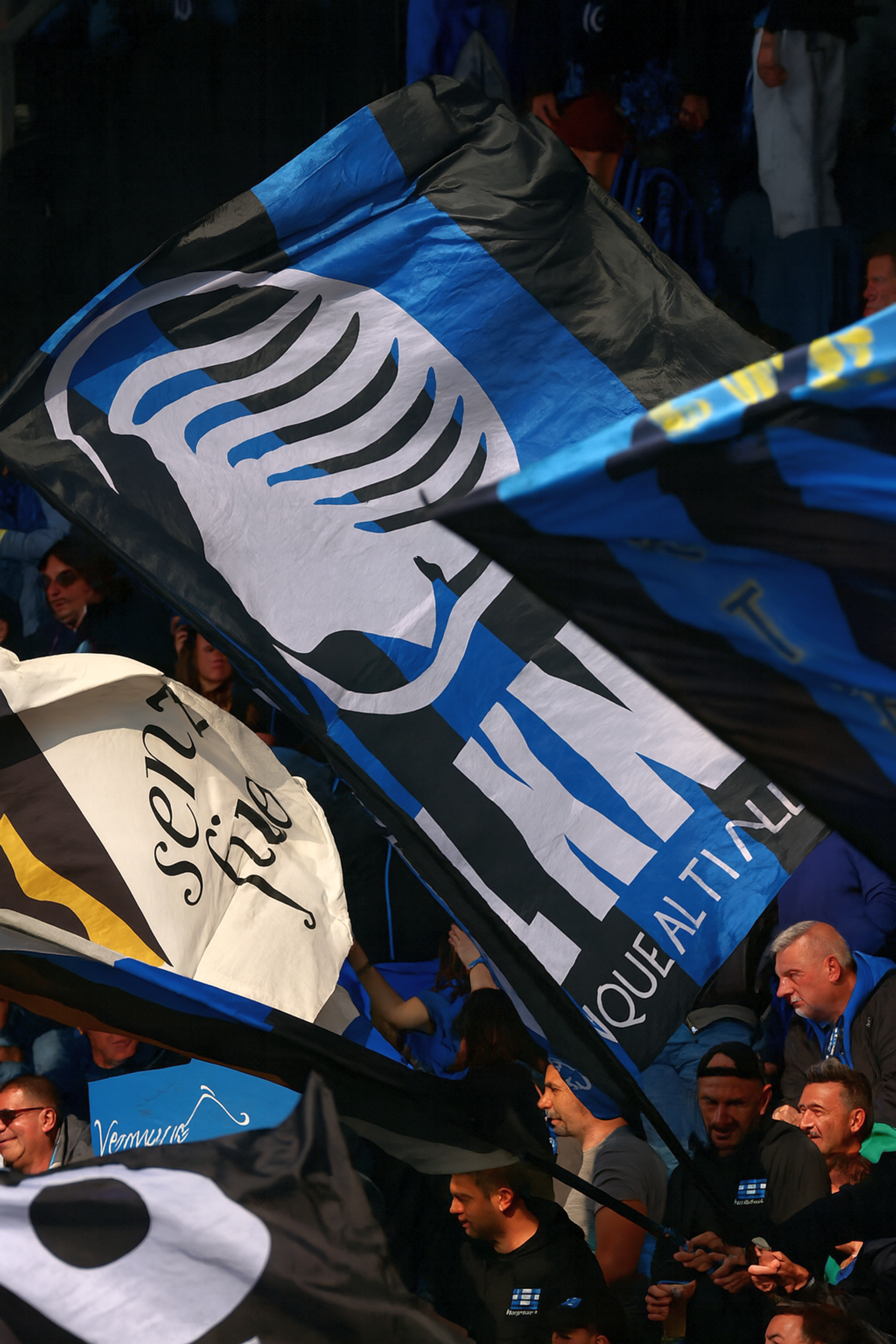 Jalkapallofanit stadionilla heiluttavat suuria mustia, sinisiä ja valkoisia lippuja, joista yhdessä on näkyvästi tyylitelty vaakuna ja sana "ATALANTA".