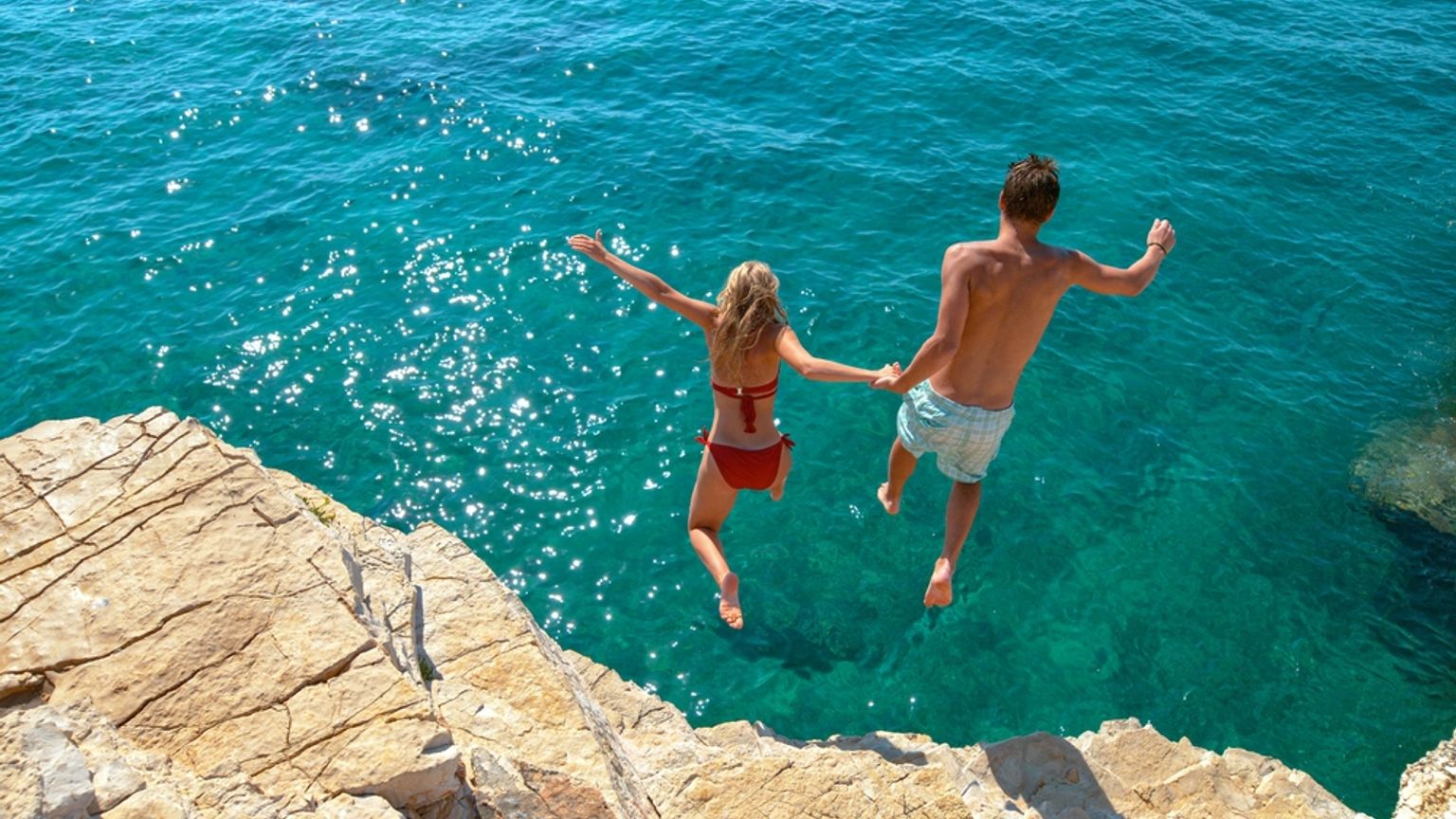 Man and woman holding hands jumping off a rocky cliff into clear blue water.