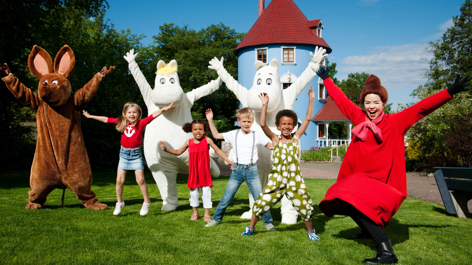 Muumi-hahmot ja lapset poseeraavat iloisesti kädet ylhäällä Muumitalon edessä.