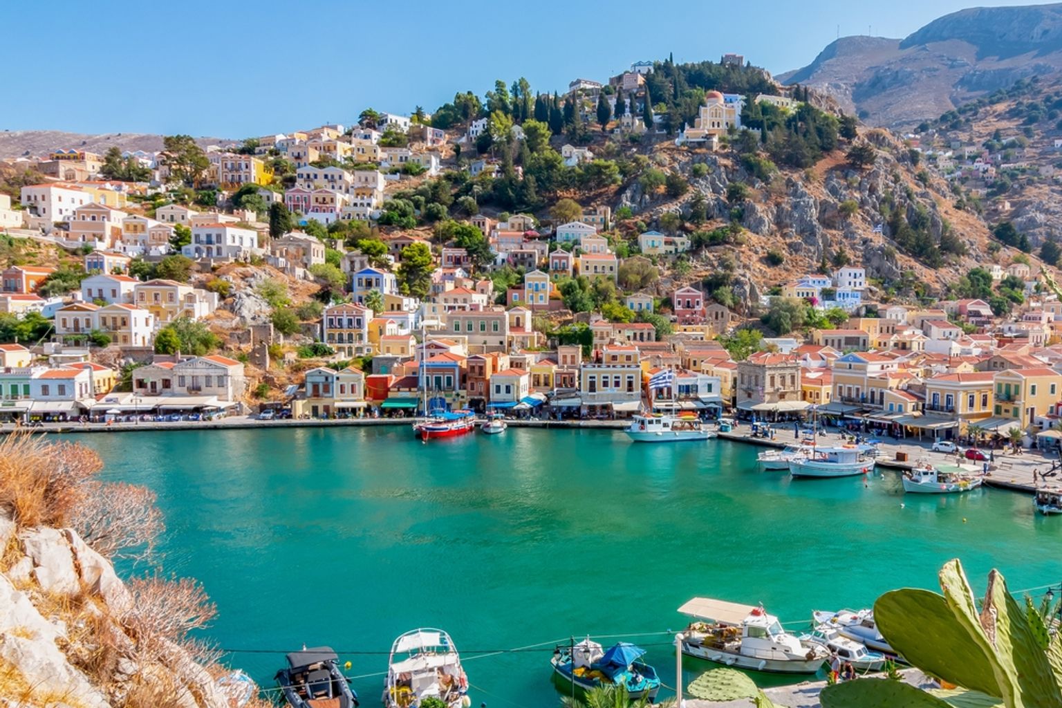 A picturesque harbor town with colorful neoclassical buildings climbing a steep hillside overlooking turquoise water filled with boats. Symi, Greece