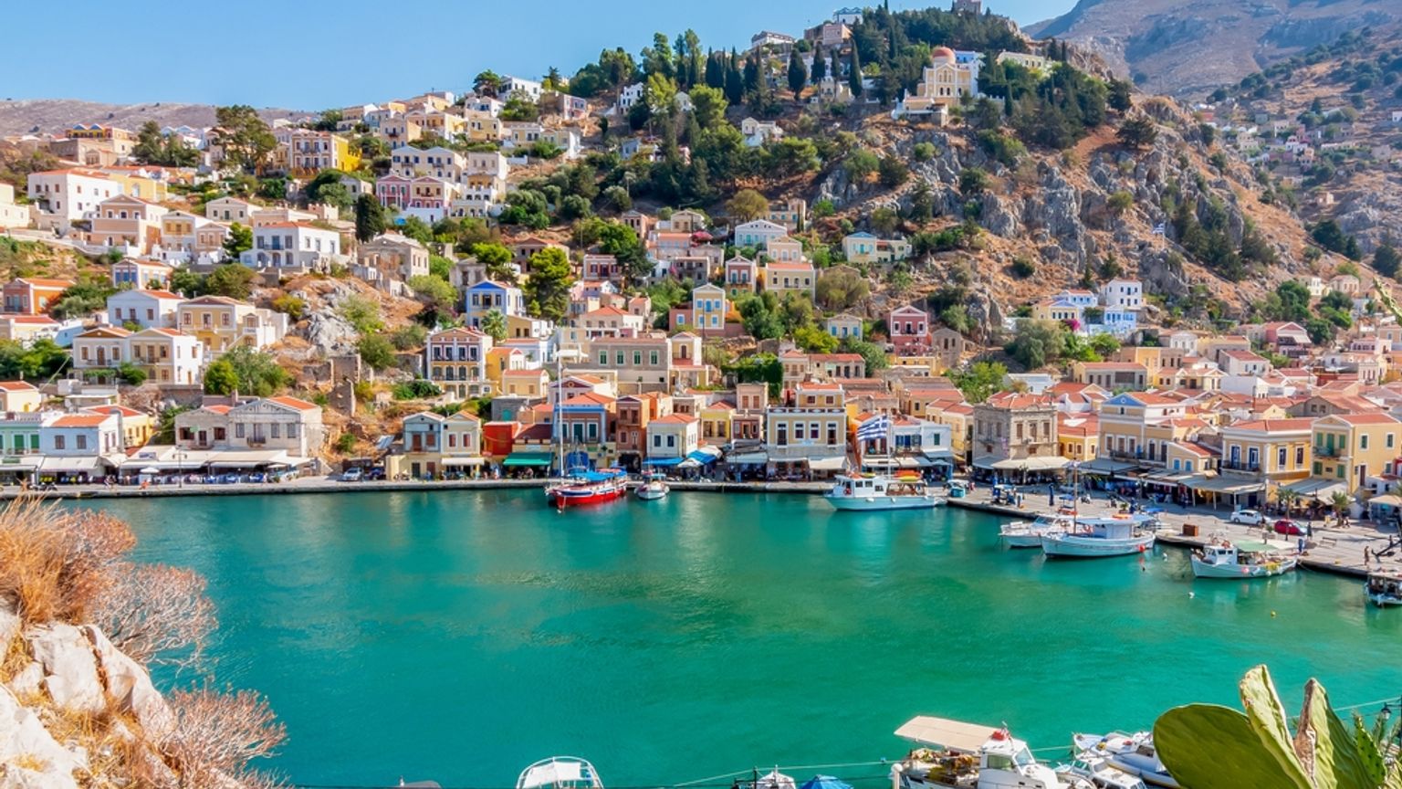 A picturesque harbor town with colorful neoclassical buildings climbing a steep hillside overlooking turquoise water filled with boats. Symi, Greece