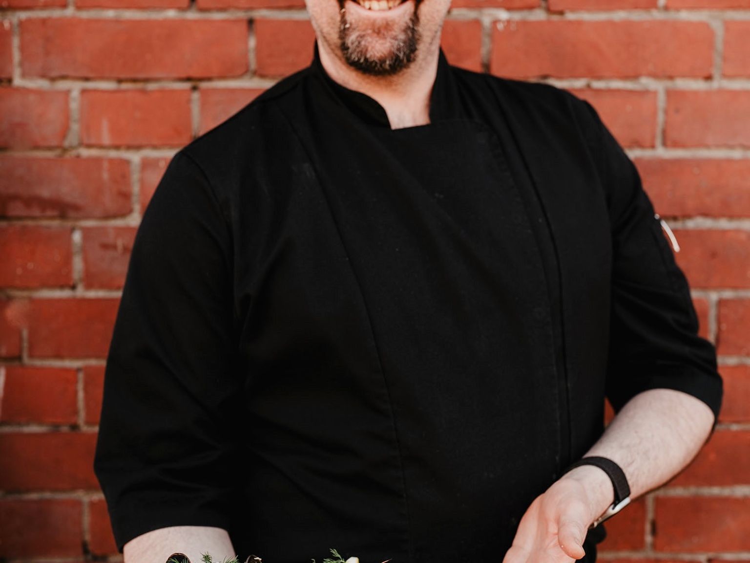 Hymyilevä kokki mustassa univormussa ja lippalakissa, joka pitää isoa jäällä olevaa merenelävälautasta.