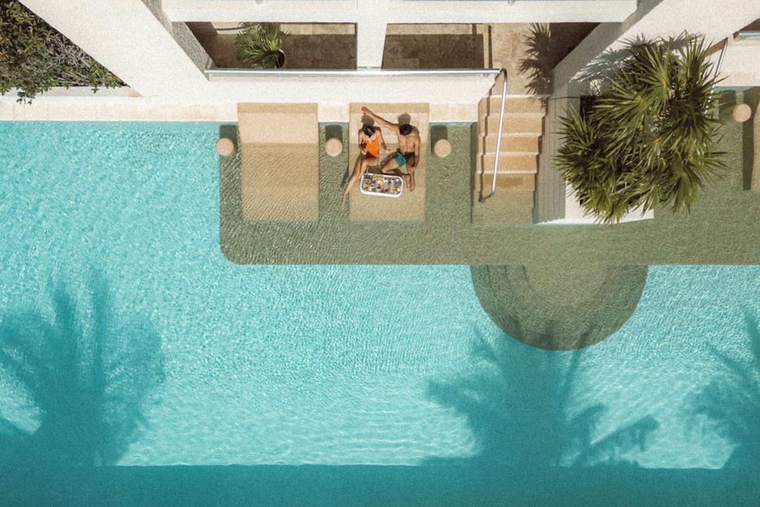 Flygvy över två personer som kopplar av på nedsänkta stolar i en pool med skuggor från palmer.