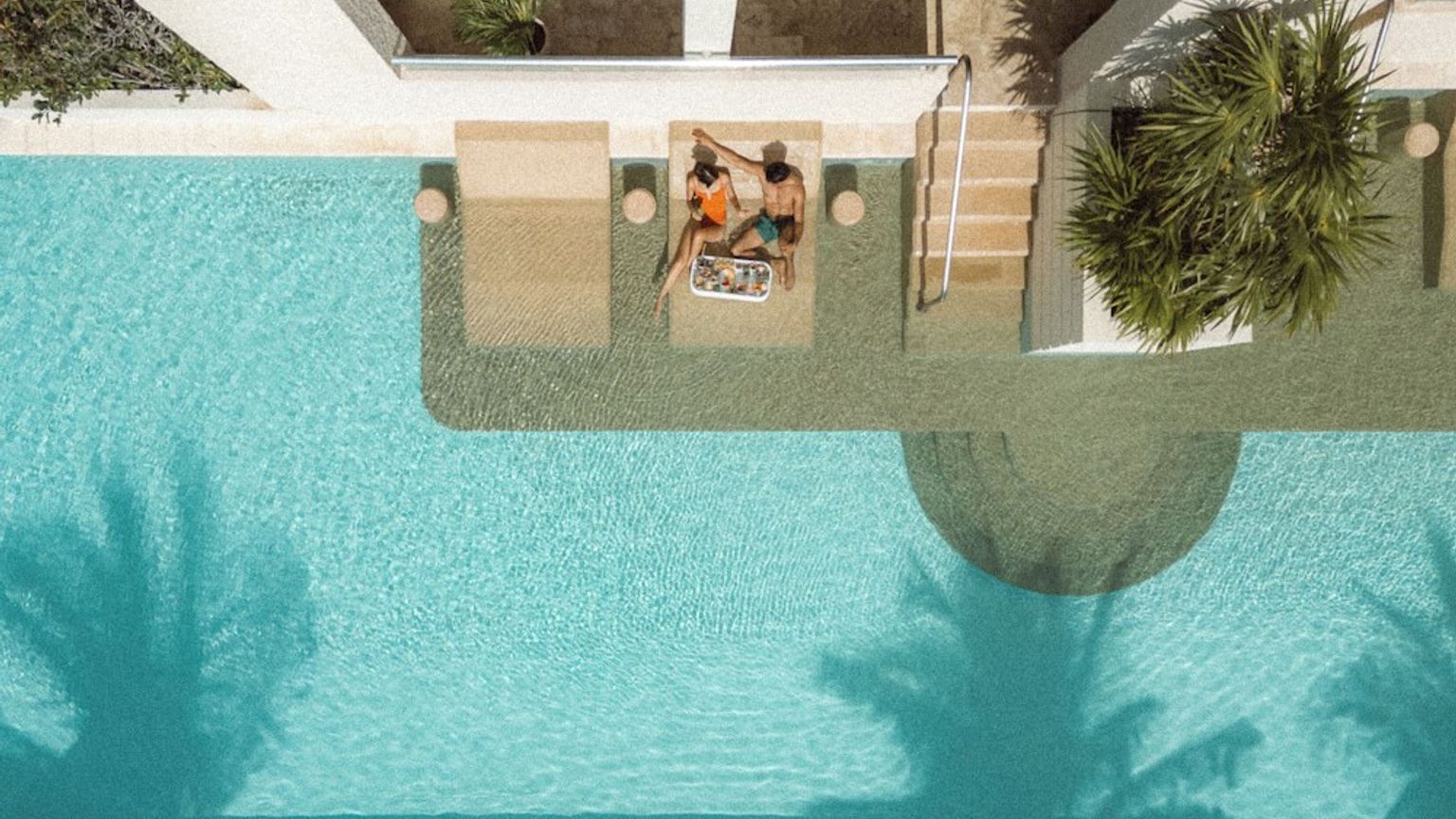 Flygvy över två personer som kopplar av på nedsänkta stolar i en pool med skuggor från palmer.